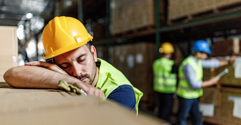 Powernapping: Ein Lagerarbeiter macht ein Nickerchen auf einem Paketstapel, Im Hintergrund stehen Kollegen am Regal. Powernapping: Ein Lagerarbeiter macht ein Nickerchen auf einem Paketstapel, Im Hintergrund stehen Kollegen am Regal.