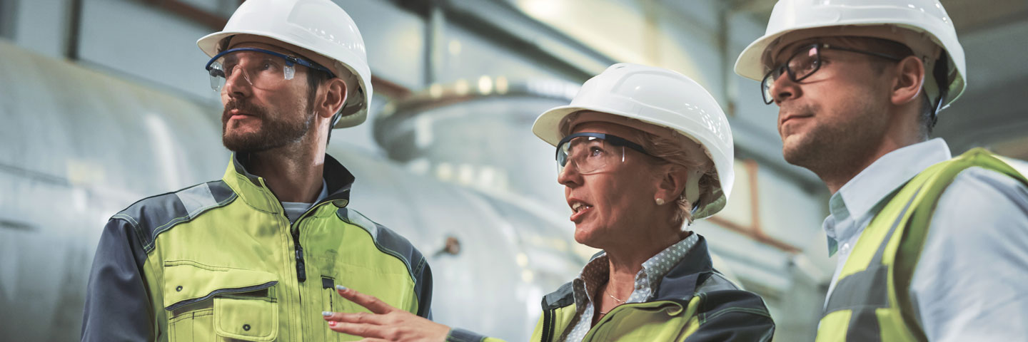 Arbeitgeber sind verpflichtet, Maßnahmen zu treffen, um den Arbeitsschutz und die Arbeitssicherheit zu gewährleisten. Zwei Männer und eine Frau in Sicherheitswesen, mit Schutzbrillen und Helmen stehen in einer Fabrikhalle.