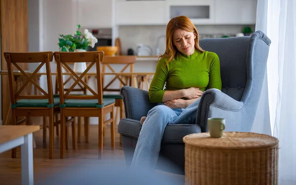 Eine junge Frau sitzt mit schmerzverzerrtem Gesicht in einem Sessel, sie hält ihr Hände vor ihrem Bauch. Im Hintergrund ist ein Esstisch mit vier Stühlen zu sehen.