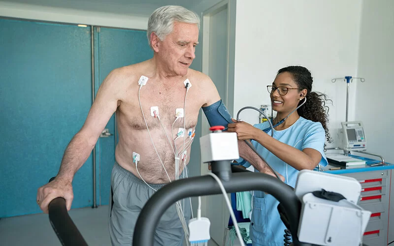 Ein älterer Mann macht gerade ein Belastungs-EKG auf einem Laufband, während eine junge Krankenschwester seinen Blutdruck kontrolliert.