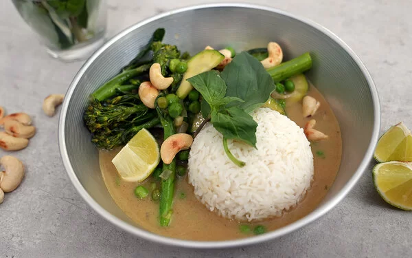 Grünes Gemüsecurry mit Zucchini nach einem Rezept von Felicitas Then, angerichtet auf einem hellgrauen Teller, garniert mit Thai-Basilikum und gerösteten Cashewkernen.