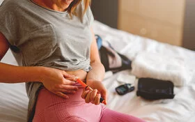 Eine junge Frau in Sportkleidung injiziert sich auf einem Bett sitzend mit einem Pen Insulin in den Bauch.