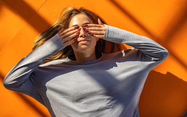 Eine junge Frau liegt rücklings auf orangefarbenem Boden und hält sich mit den Händen die Augen zu.