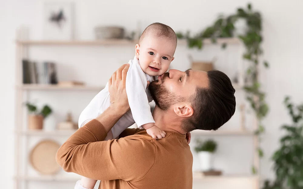 Ein junger Vater hebt in seinem Wohnzimmer sein Baby hoch und küsst es auf die Wange.