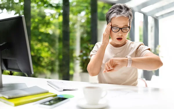 Eine Frau mit leicht grauen Haaren und Brille sitzt an einem Schreibtisch, vor sich Monitor, Tastatur und einige Unterlagen. Sie blickt auf ihre Armbanduhr, fasst sich an die Stirn und schaut erschrocken.
