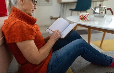 Eine ältere Frau mit dunkler Brille und orangefarbenem kurzärmeligen Rollkragenpullover, die vor einem Couchtisch am Boden sitzt, schreibt etwas in ein Notizbuch.