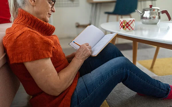 Eine ältere Frau mit dunkler Brille und orangefarbenem kurzärmeligen Rollkragenpullover, die vor einem Couchtisch am Boden sitzt, schreibt etwas in ein Notizbuch.