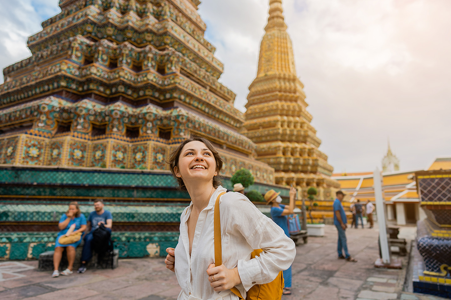 Eine Frau mit Rucksack steht lächelnd vor dem kunstvoll verzierten Tempel Wat Pho in Bangkok und schaut nach oben.
